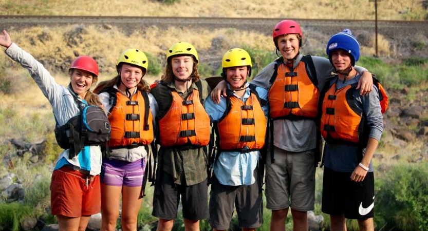Six people wearing helmets and life jackets stand on land, wrap their arms around each other and smile at the camera.